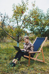 Naklejka na ściany i meble A cute young female freelancer is working in the garden outdoors while sitting in a comfortable garden chair. Remote work on a portable laptop