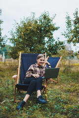 Stylish woman sitting on a chair near green plants in the garden and browsing the internet on a laptop while looking to the side. Remote work, female freelancer