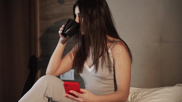 Happy smiling woman using smartphone and dinking hot coffee in the morning on bed Young woman in bed with smart phone and cup of coffee in hand. Happy female using mobile phone and internet