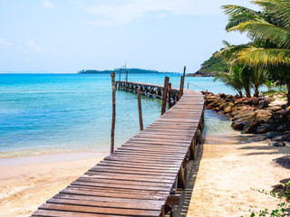 Wooden bridge heading to the blue sea. Brown wood plank pathway bridge on the beach sand with rocks and coconut palm tree at the local port in island on sunny day. Seascape summer holiday background.