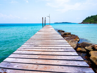 Wooden bridge heading to the blue sea. Brown wood plank pathway bridge on the beach sand with rocks at the local port in the island on sunny day. Seascape summer holiday background.