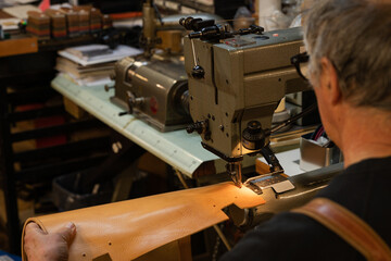 view of a leather craftsman’s shoulders sitting while sewing a leather back of a chair in his traditional workshop in the historic center of rome, italy