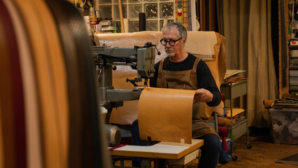 senior tanner sitting sewing leather back panel with a sewing machine for a chair made by him in workshop