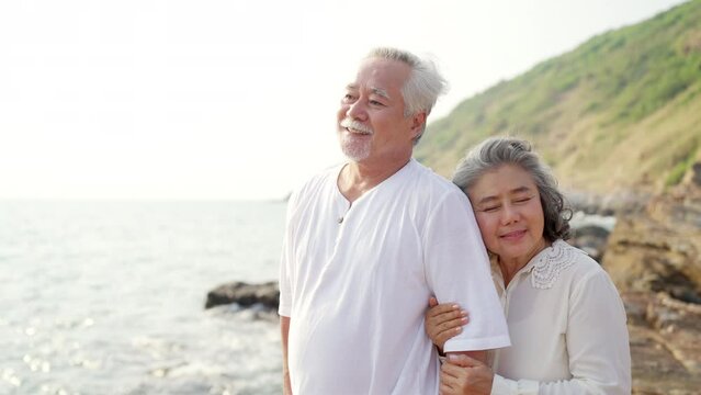 Happy Asian family senior couple with gray hair relaxing and hugging each other on mountain cliff at sunset. Retirement elderly people enjoy outdoor lifestyle travel nature ocean on summer vacation.