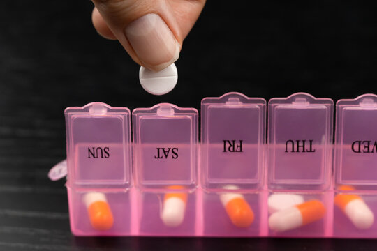 Close-up of a woman with her hand taking pills from a pink pillbox or sorting pills and vitamins for daily medication on a black table. Concept of healthcare, medicine