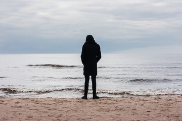 depressed man in black clothes with a hood stands by the sea in the sand on the shore and looks into the distance.