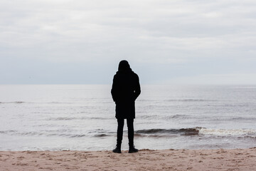 depressed man in black clothes with a hood stands by the sea in the sand on the shore and looks into the distance.