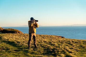 Bearded Man reaching the destination  at sunset on cool day and taking photos of amazing seaside landscape in Wales. Travel Lifestyle concept.
