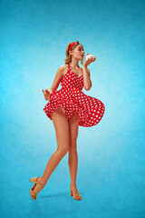 Sweet tooth. Full-length portrait of young pretty blonde girl in stylish red dress posing against blue background, eating cake. Concept of retro fashion, beauty, 50s, 60s. Pin-up style