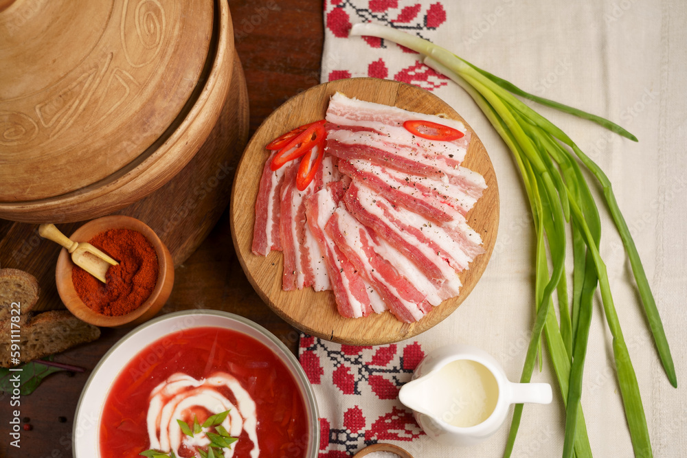 Poster Pieces of salted fatty meat cooked with spices. Salo, bacon, ham. Garlic, fresh onion, spices. A plate of Ukrainian borscht. wooden background