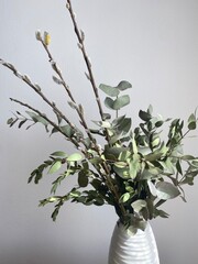 A bouquet of green eucalyptus and pistachio branches with willow branches in a white vase on a chest of drawers. Natural elements of spring