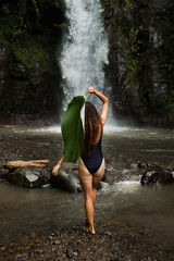 A beautiful girl in a swimsuit with a palm leaf stands in front of a waterfall, rear view.
