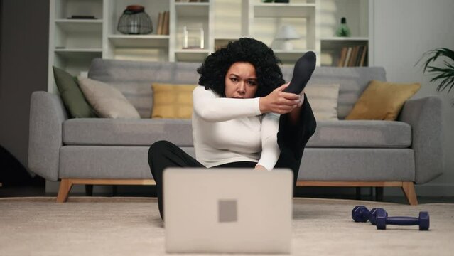 Funny african american young woman with puzzled face looks at hard fitness sport workout of online trainer and unsuccessfully tries to repeat at home Curly female beginner makes a clumsy move indoors