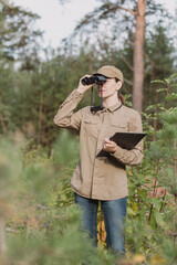 A woman park ranger in uniform looks through binoculars at the forest area with a clipboard in hand...