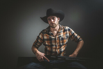 Rugged man with beard and a shotgun in cowboy hat.  Plaid shirt. Blue jeans. Studio session.
