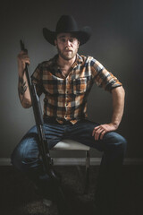 Rugged man with beard and a shotgun in cowboy hat.  Plaid shirt. Blue jeans. Studio session.