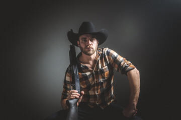 Rugged man with beard and a shotgun in cowboy hat.  Plaid shirt. Blue jeans. Studio session.