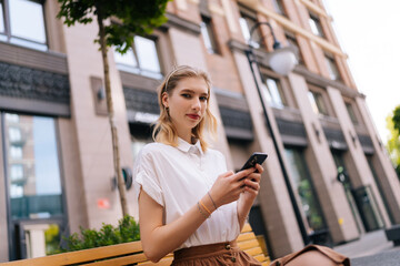 Low-angle view of attractive female blonde young woman sitting on bench on city street and looking at camera holding smartphone in hand on background of building. Lady using mobile phone outdoors.