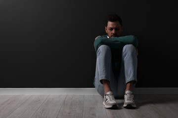 Upset man sitting on floor near black wall. Space for text