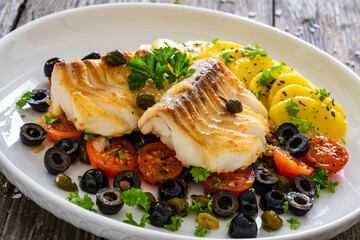 Cod in Italian style on tomatoes and black olives with baked potatoes  on wooden table