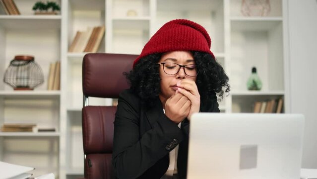 Cold curly african american businesswoman in red hat trembles warms hands try to work on laptop computer at desk in office workplace indoors Problem of heating system or low temperature concept