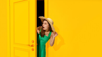 Summer vacation. Young emotive woman in dress and straw hat peeking out yellow door with shocked, surprised, astonished face. Concept of emotions, facial expression, lifestyle