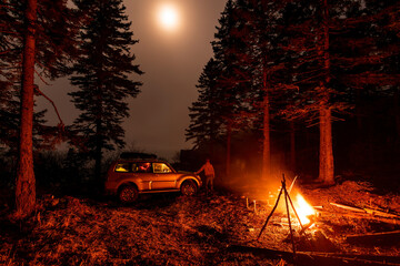 A man stands at his car in a tourist camp in a fir forest near the sea.
