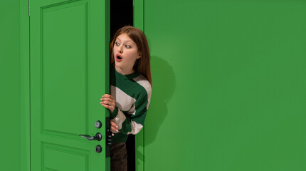 Astonished young girl, teengager peeking out green door with shocked face, looking with wide open...