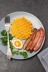 Waffles, fried eggs, bacon and a fork on a gray plate.