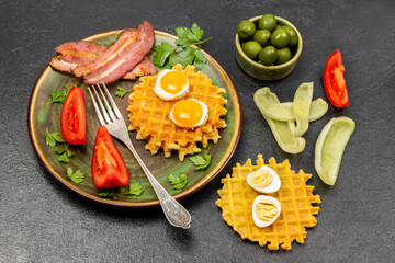 Fototapeta na wymiar Waffles, fried eggs, bacon and fork on plate. Olives in bowl. Chopped vegetables and waffles on table.