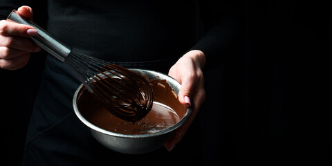 A bowl of melted chocolate and a kitchen whisk are held by hands. Kitchen utensils. On a black...
