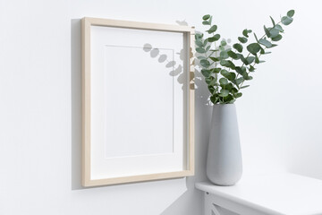 Portrait wooden picture frame mockup in white room interior with fresh eucalyptus twigs, blank frame with copy space