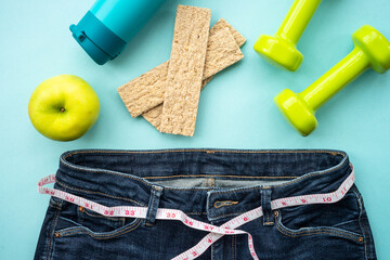 Losing weight background. Dumbells, healthy food and jeans with measuring tape. Flat lay on blue.