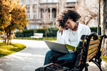 Beautiful and serious African female student enjoying outdoors in college campus while study for the faculty exam. She is sitting on park bench and reading something on her laptop computer.
