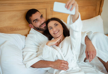 Obraz na płótnie Canvas Happy Couple Using Smartphone Making Selfie Posing Lying In Bedroom