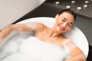 Naklejka premium Female Relaxing Listening Music Wearing Earbuds In Bathroom, Above View