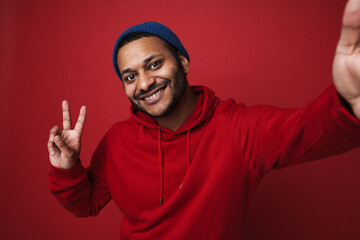 Smiling indian man taking selfie and showing peace sign isolated over red background