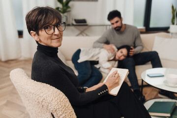 Psychologist writing down notes and smiling at camera during therapy session with couple