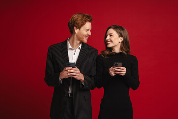 Cheerful couple using mobile phones while standing isolated over red wall