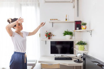 Young woman adjusts the temperature of the air conditioner using the remote control in room at home - obrazy, fototapety, plakaty