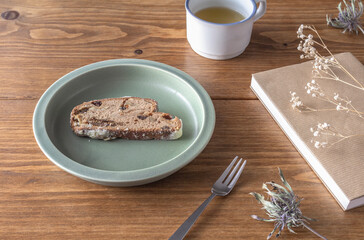 A round khaki plate with baked goods placed randomly on a brown wooden table, a fork, a book, dried flowers, and tea.