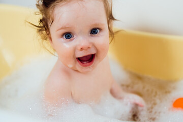 Baby girl in a bath with foam and soap bubbles. Happy laughing baby taking a bath playing with toys. Little child in a bathtub. Smiling kid in bathroom. Infant washing and bathing