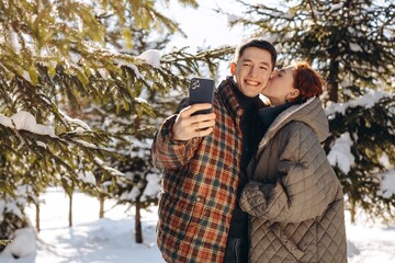 A young man takes a selfie as a girl kisses him on the cheek. A young married couple walking in a winter park makes a joint photo for social networks.