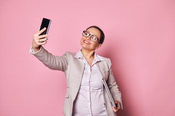 Attractive middle-aged multi-ethnic pregnant woman, business person wearing casual clothes and trendy glasses, carrying laptop and taking selfie on her smartphone on isolated pink background. Ad space