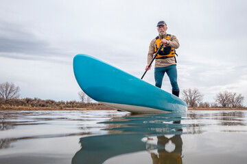 solo senior male paddling a stand up paddleboard on a lake in early spring, frog perspective from an action camera at water level - obrazy, fototapety, plakaty