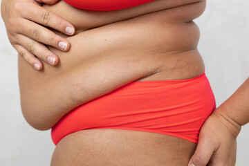 Cropped close up profile of fat, overweight big woman in red bikini touch and hold sagging flabs sides on belly on white background. Subcutaneous fat and superfluity of calories. Need abdominoplasty