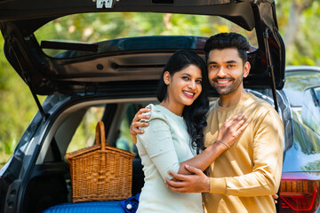 Happy young couple in front of car with travel bags and picnic basket standing by embracing each...