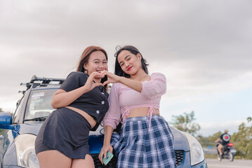 Fototapeta na wymiar Two women at the side of the road, side to side making a heart shape sign with each others hands smiling widely while looking at the camera and leaning on their blue car