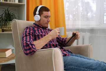 Obraz premium man in white headphones and a vibrant red checkered shirt, sitting in a cozy chair with a cup of coffee in hand, lost in the world of his smartphone. taking a break from the daily grind