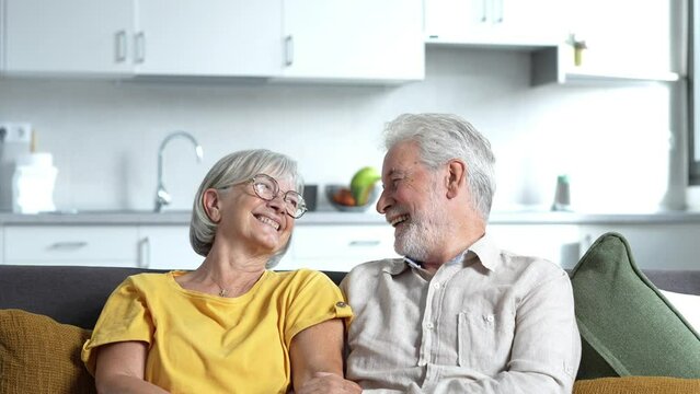 Headshot portrait of smiling elderly 60s husband and wife sit relax on couch hugging cuddling, happy mature old couple rest on sofa in living room embrace look at camera show love and care
